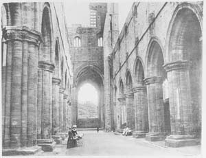 Black and white photograph looking down the nave of Kirkstall Abbey after the programme of conservation in 1892-6. The abbey re-opened as a public park in 1895 and here we can see Victorian or Edwardian visitors exploring the ruins.