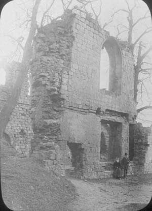 The north end of the refectory at Kirkstall Abbey showing a large pile of debris from the collapse of he buildings and large elm trees growing close to the walls. Two people in Victorian dress can be seen on the right. Large elm trees are growing in the debris inside the building. The photograph must have been taken, therefore, before 1892-6.